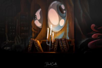 Un chandelier allumé éclaire des livres anciens posés sur un pupitre en bois, devant une fresque sombre et colorée.