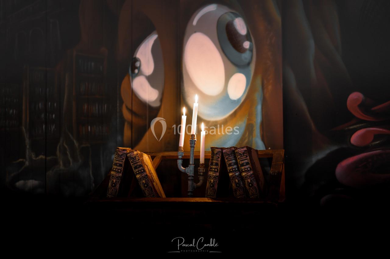 Un chandelier allumé éclaire des livres anciens posés sur un pupitre en bois, devant une fresque sombre et colorée.