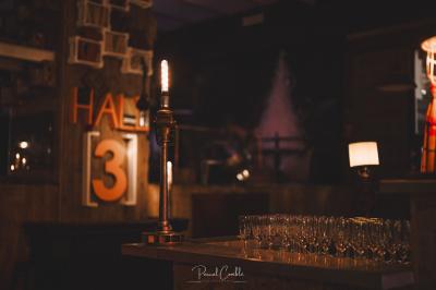 Un chandelier allumé éclaire des livres anciens posés sur un pupitre en bois, devant une fresque sombre et colorée.