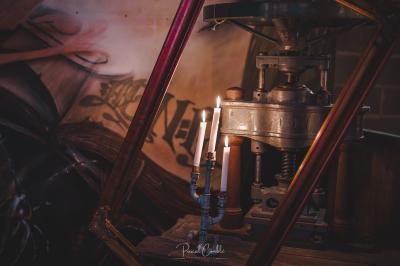 Un chandelier allumé éclaire des livres anciens posés sur un pupitre en bois, devant une fresque sombre et colorée.