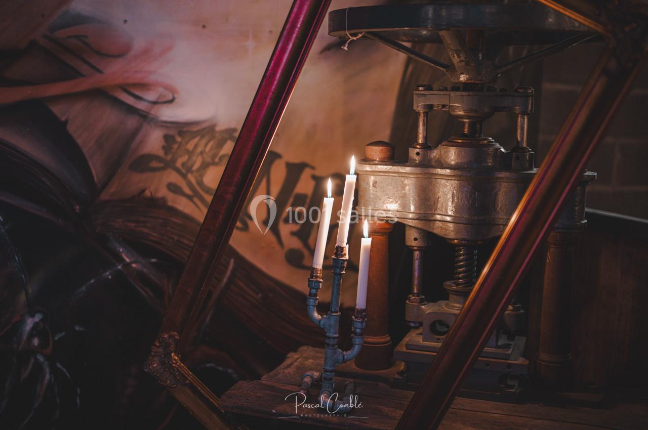 Chandelier à bougies allumées posé près d'une ancienne presse métallique, avec une fresque en arrière-plan.