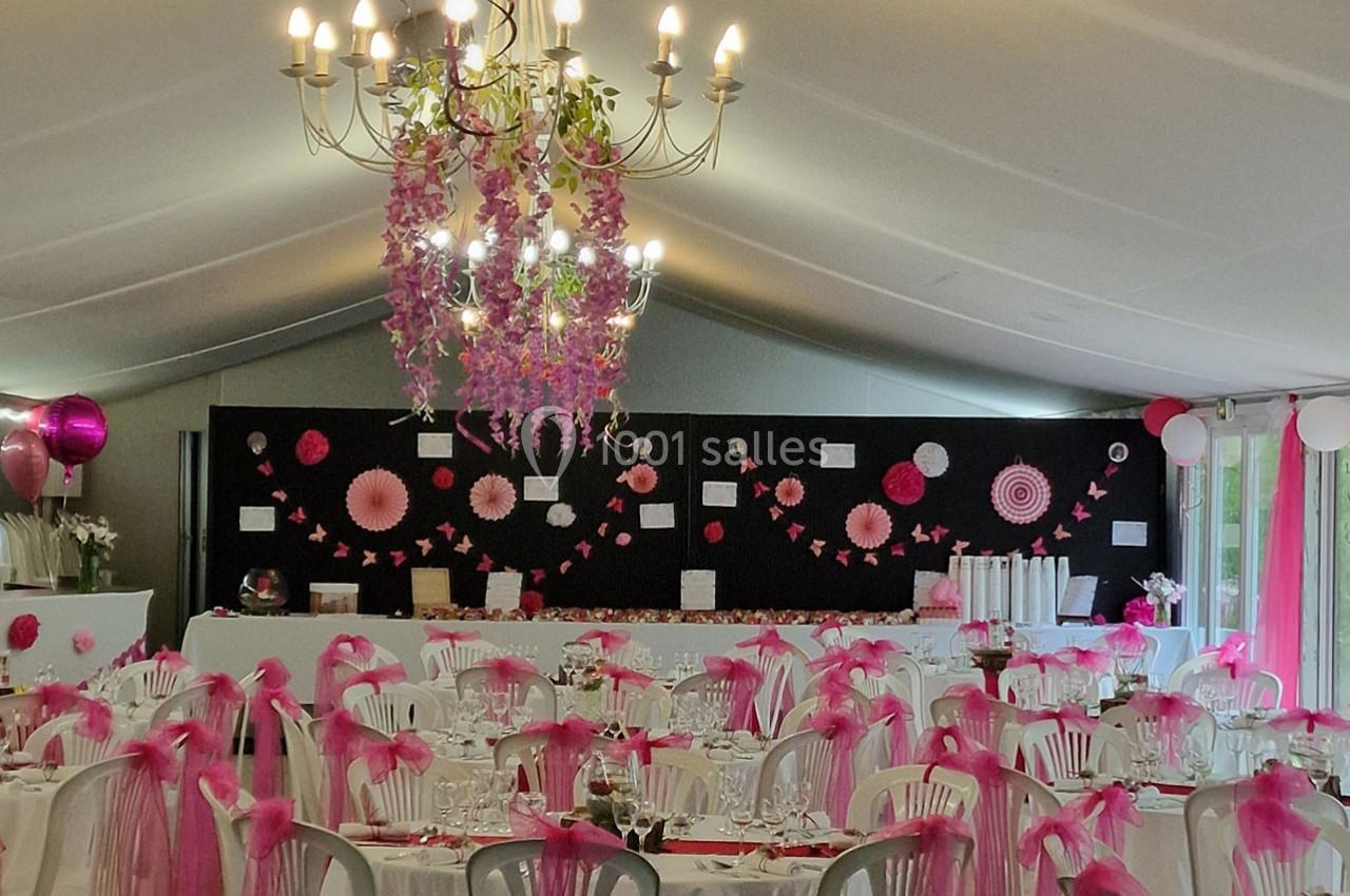 Salle de réception décorée en rose et blanc avec des guirlandes, des fleurs suspendues et des tables dressées.
