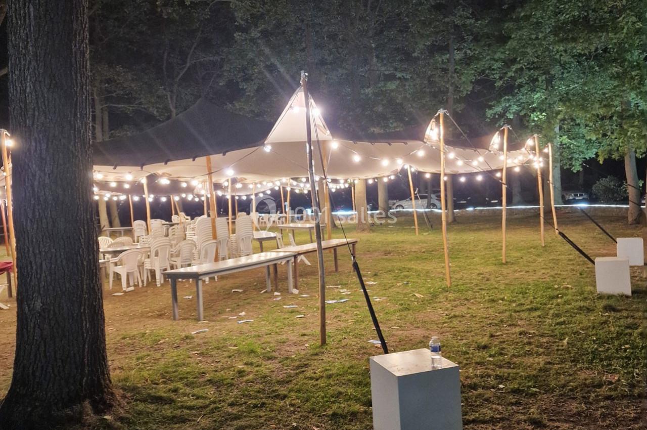 Tente éclairée par des guirlandes lumineuses dans un espace extérieur, avec des chaises et tables en plastique disposées.