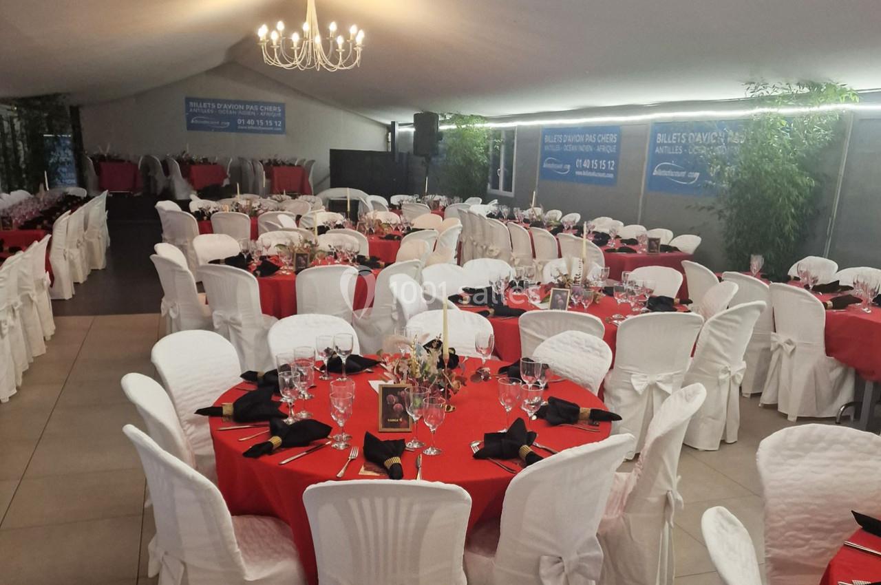 Salle de réception avec tables rondes dressées, nappes rouges, chaises blanches et décoration élégante.