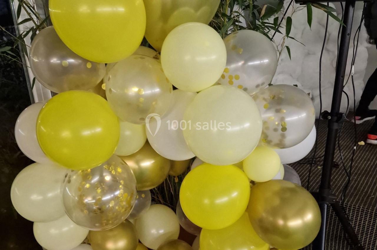 Bouquet de ballons jaunes, blancs et dorés avec des motifs de confettis, disposé devant des plantes vertes.