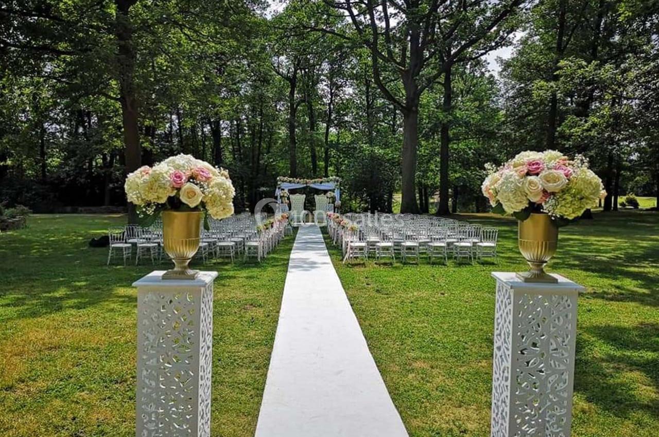 Allée centrale décorée de fleurs menant à une arche de mariage dans un jardin verdoyant avec des chaises alignées.