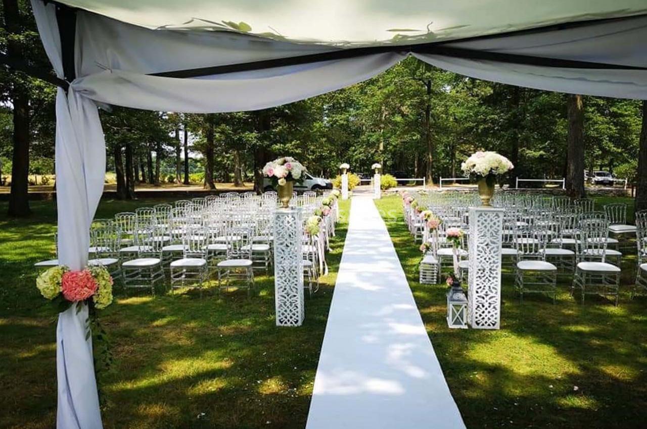 Allée centrale décorée pour une cérémonie en plein air, entourée de chaises et de fleurs sous un auvent blanc.