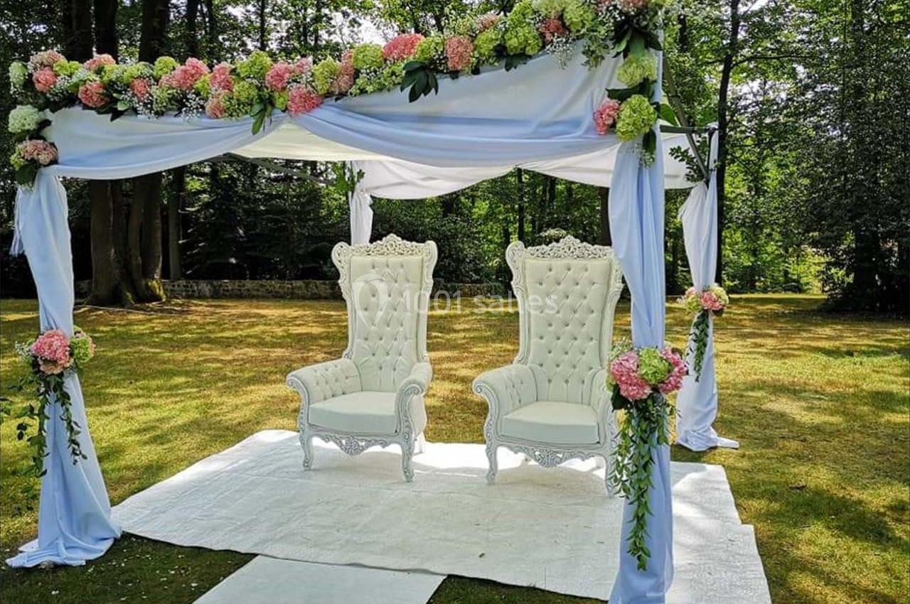 Deux fauteuils blancs ornés de détails sculptés sous une arche décorée de fleurs et de tissus dans un jardin.