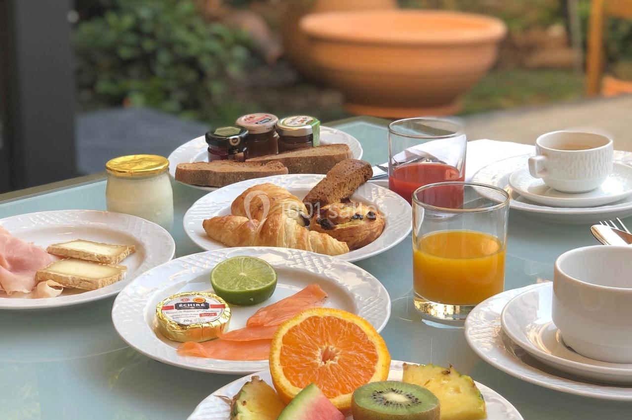 Petit-déjeuner varié avec fruits, viennoiseries, saumon, charcuterie, confitures et boissons sur une table en extérieur.