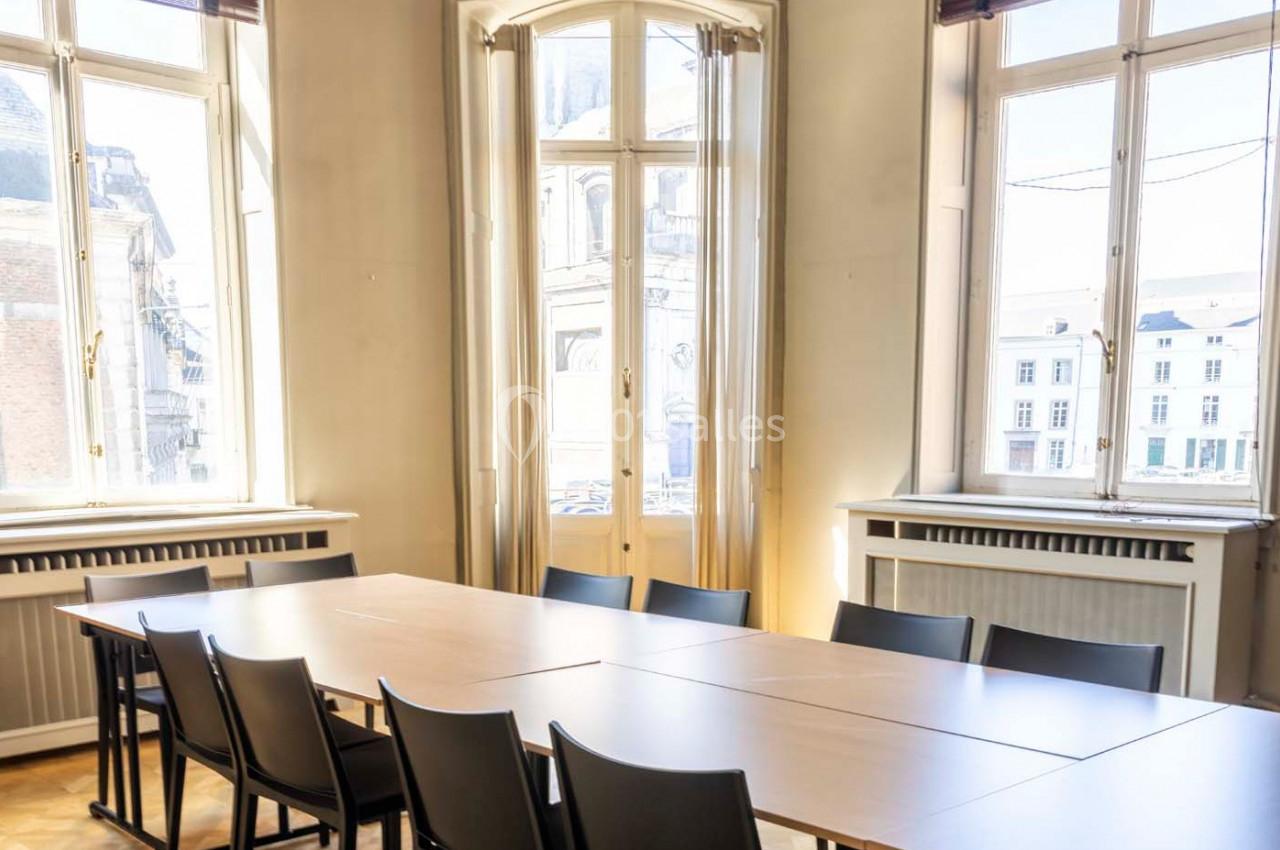 Salle de réunion lumineuse avec une grande table en bois entourée de chaises, près de fenêtres hautes.