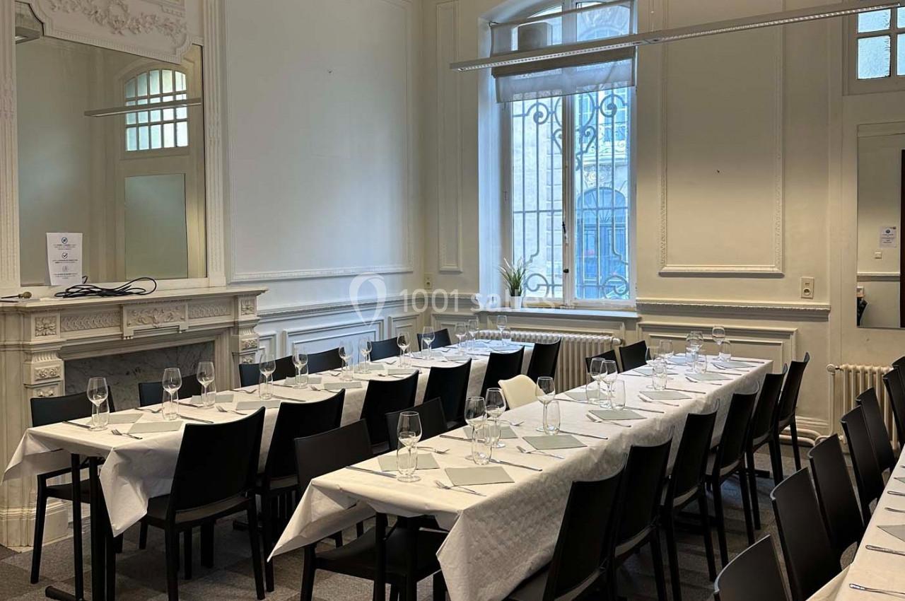 Salle de réception lumineuse avec tables alignées, nappes blanches, verres et chaises noires, décor sobre et élégant.