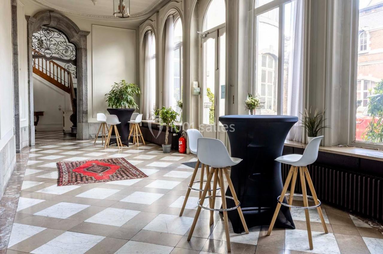 Salle lumineuse avec grandes fenêtres, sol en carreaux, tables hautes, chaises blanches et plantes décoratives.
