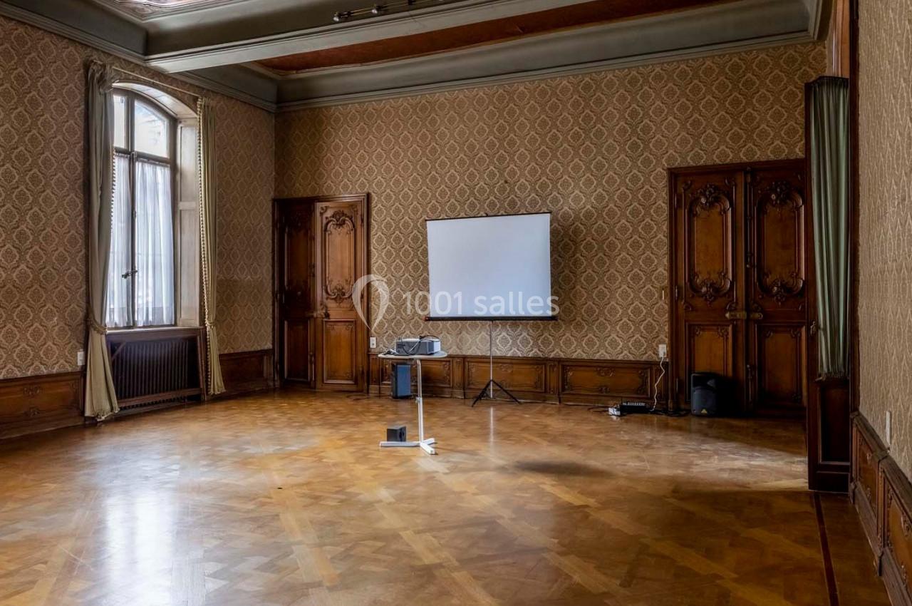 Salle vide avec parquet, murs ornés de motifs, grandes fenêtres, rideaux et un écran de projection installé.