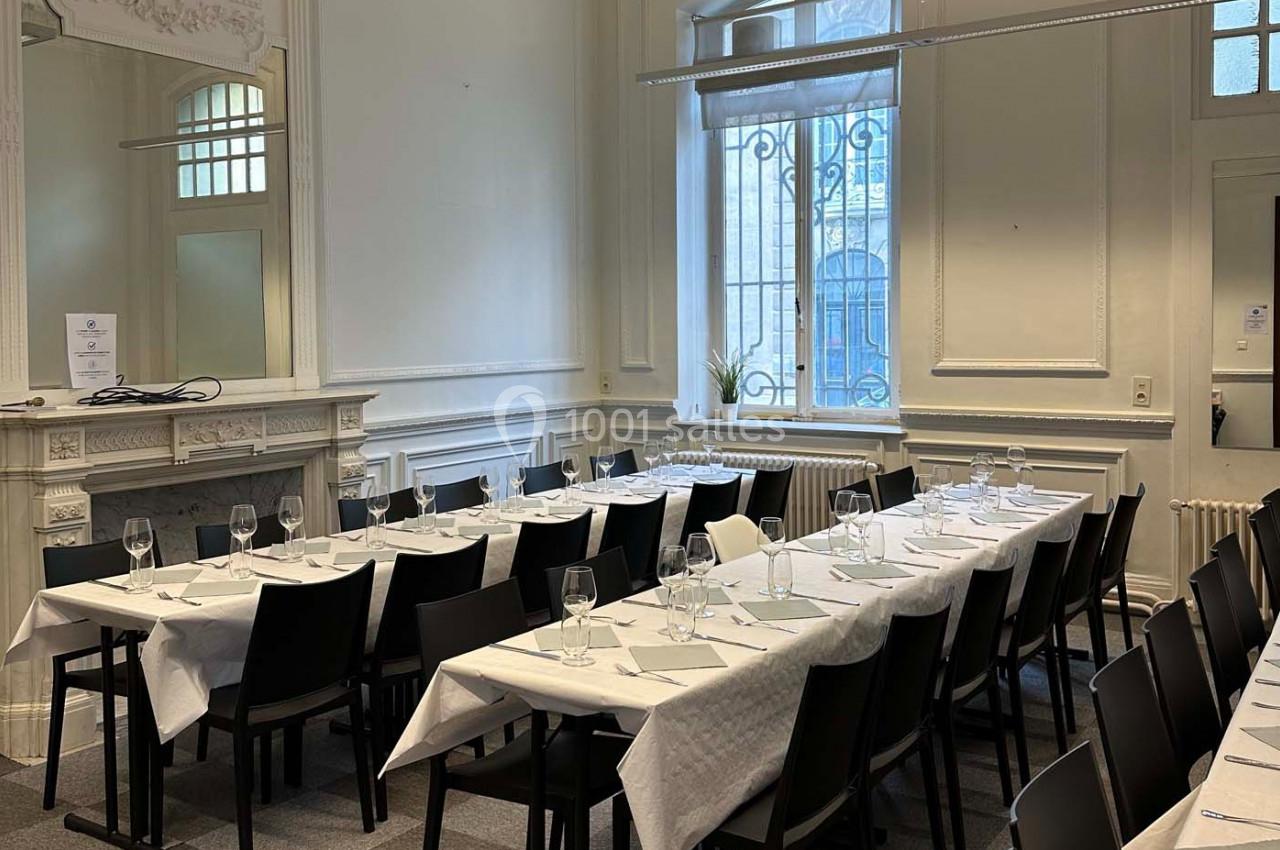 Salle lumineuse avec tables alignées, nappes blanches, verres et couverts, décorée de moulures et d'une cheminée.