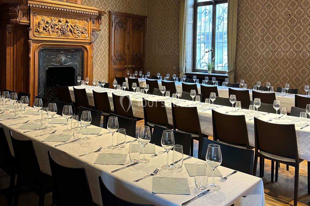 Salle élégante avec tables alignées, nappes blanches, couverts et verres, décorée de boiseries et d'une cheminée sculptée.