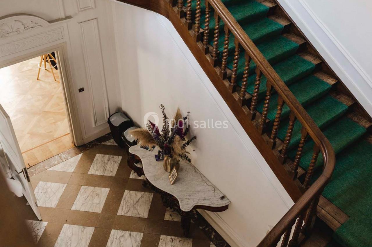 Vue d'un hall d'entrée avec un escalier en bois, tapis vert, table en marbre et bouquet de fleurs séchées.