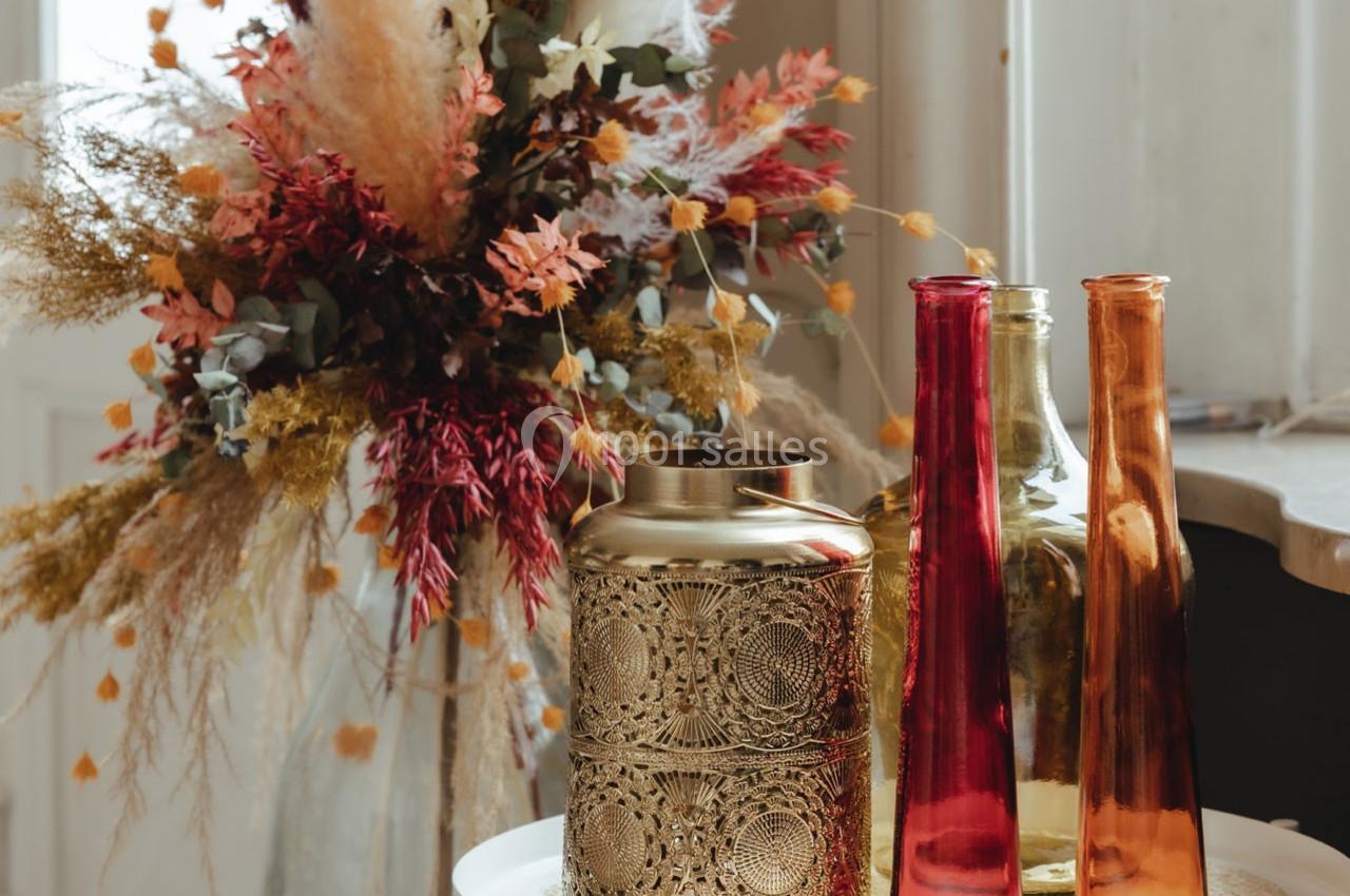Vases colorés et doré décoratif posés sur une table, avec un bouquet de fleurs séchées en arrière-plan.