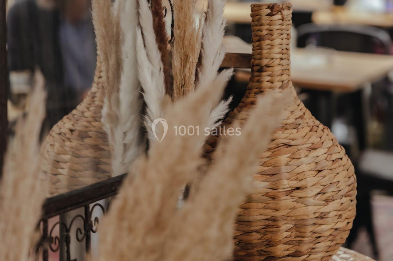 Vase en osier tressé entouré de pampas séchées dans un décor intérieur chaleureux avec des tables en arrière-plan.