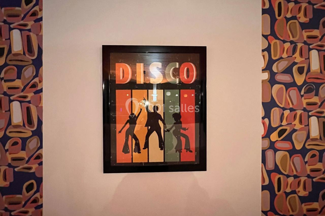 Illustration encadrée représentant des silhouettes dansantes sous le mot ’DISCO’, accrochée sur un mur à motifs colorés.