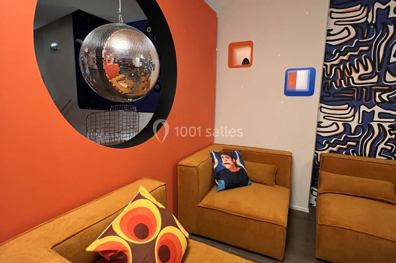 Salon moderne avec fauteuils en velours orange, coussins colorés, boule à facettes et décoration murale graphique.