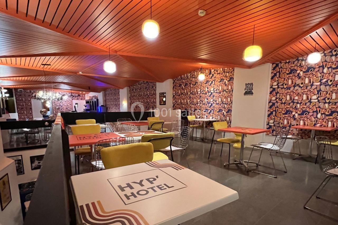 Salle de restaurant moderne avec tables colorées, chaises variées et plafond en bois éclairé par des suspensions.