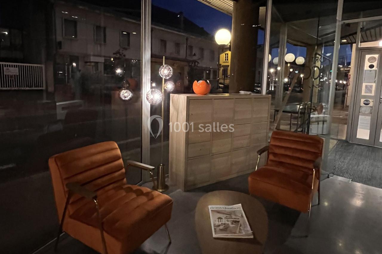 Deux fauteuils orange et une table basse avec un journal dans un espace vitré éclairé par des lampes en soirée.