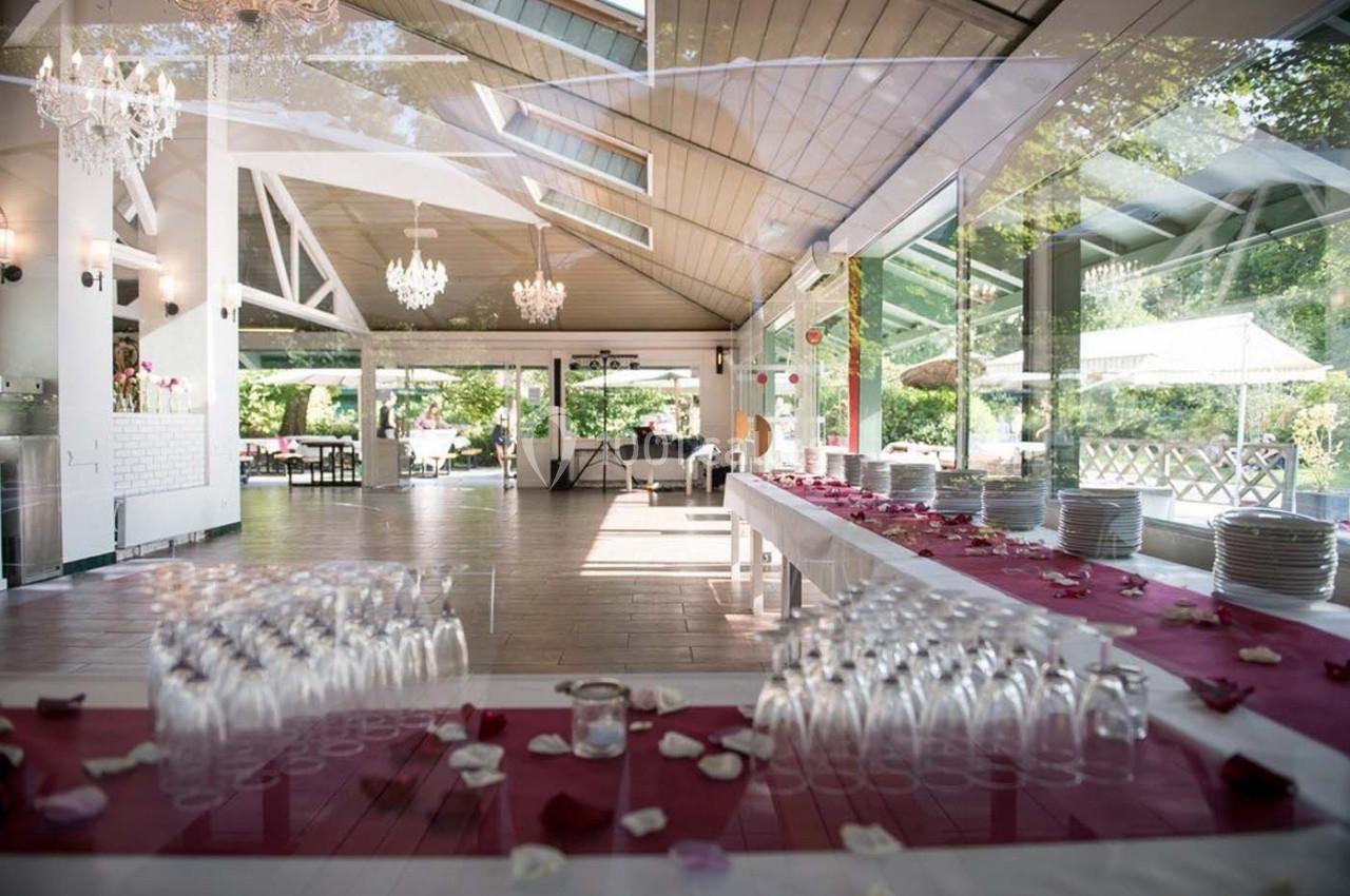 Salle lumineuse avec de grandes baies vitrées, tables dressées avec nappes rouges, pétales et verres alignés.