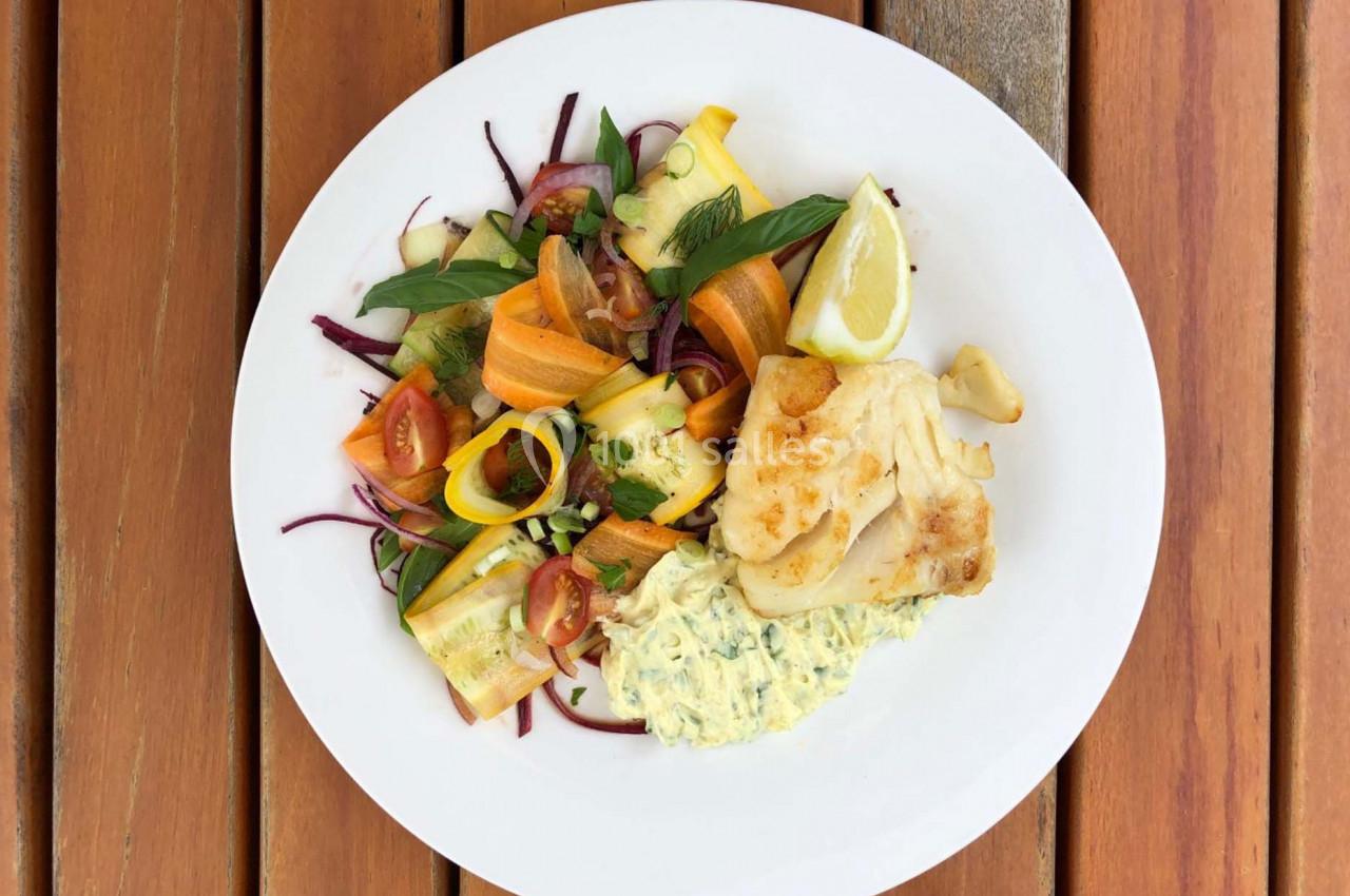 Assiette contenant du poisson grillé, salade de légumes colorés et sauce, servie sur une table en bois.