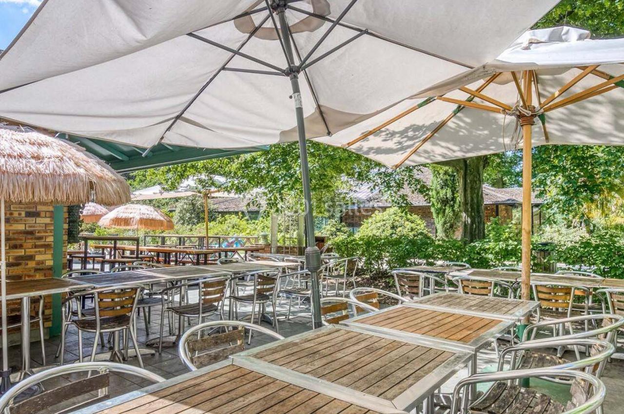 Terrasse extérieure avec tables et chaises en bois et métal, ombragée par des parasols et entourée de verdure.