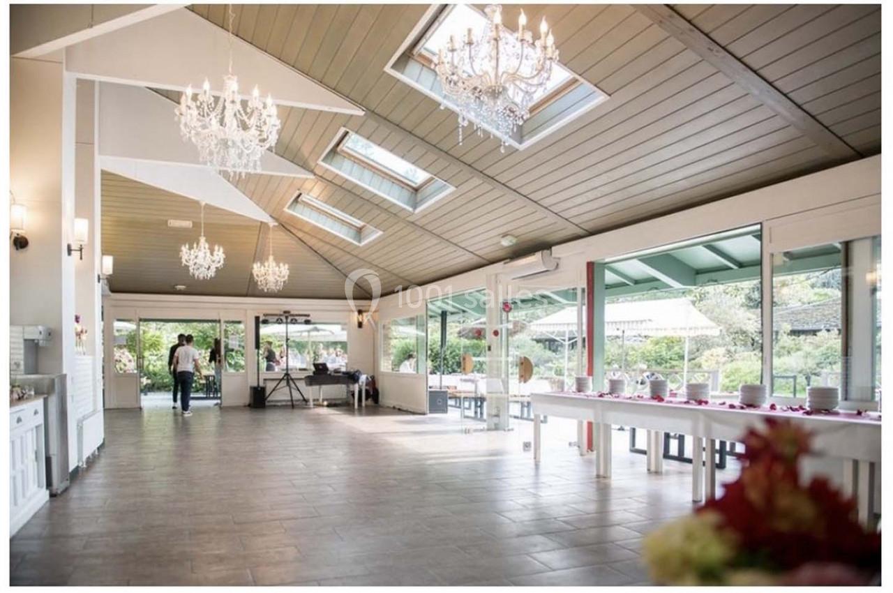 Salle lumineuse avec baies vitrées, tables dressées et lustres suspendus, donnant sur un espace extérieur verdoyant.