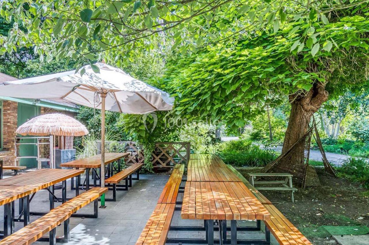 Tables en bois sous des parasols dans un jardin ombragé, entouré de verdure et d'arbres.
