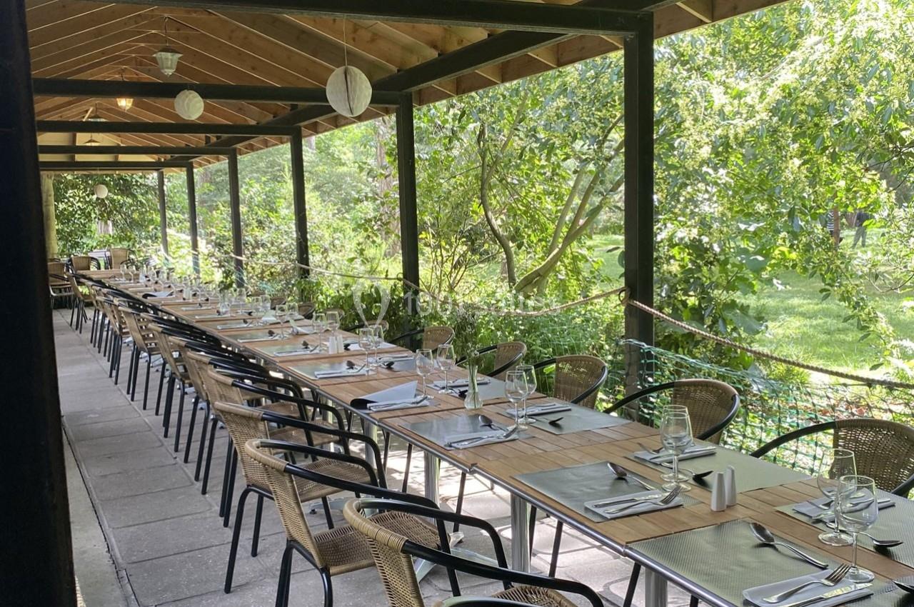 Terrasse couverte avec de longues tables dressées, entourée de verdure et éclairée par des lanternes suspendues.