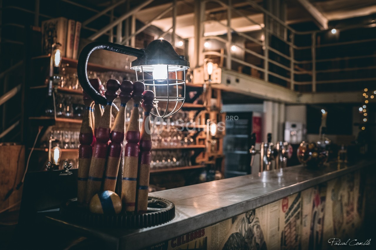Lampe industrielle éclairant un comptoir en métal, décoré de quilles et entouré d'étagères remplies de verres.