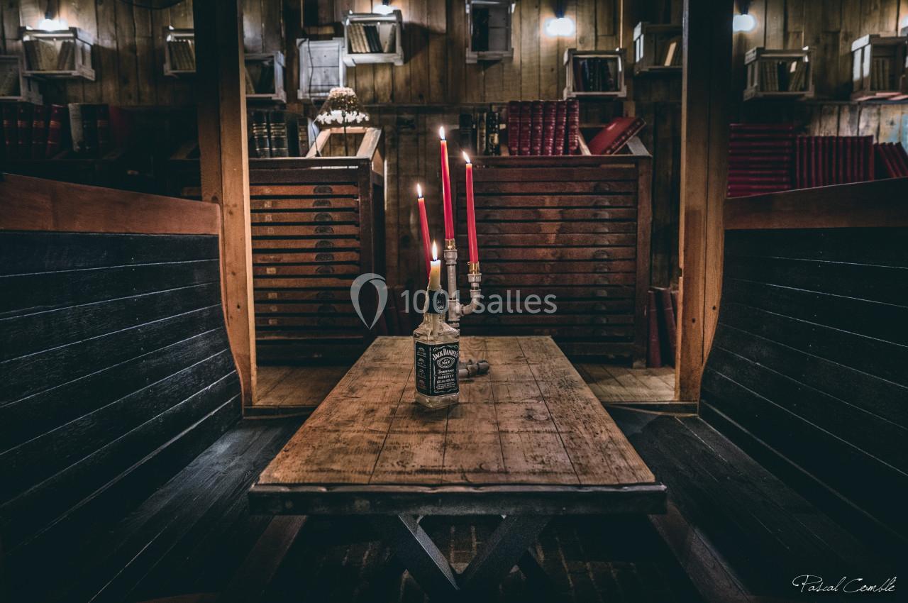 Table en bois avec une bouteille transformée en chandelier à trois bougies rouges, dans un intérieur rustique avec étagères.