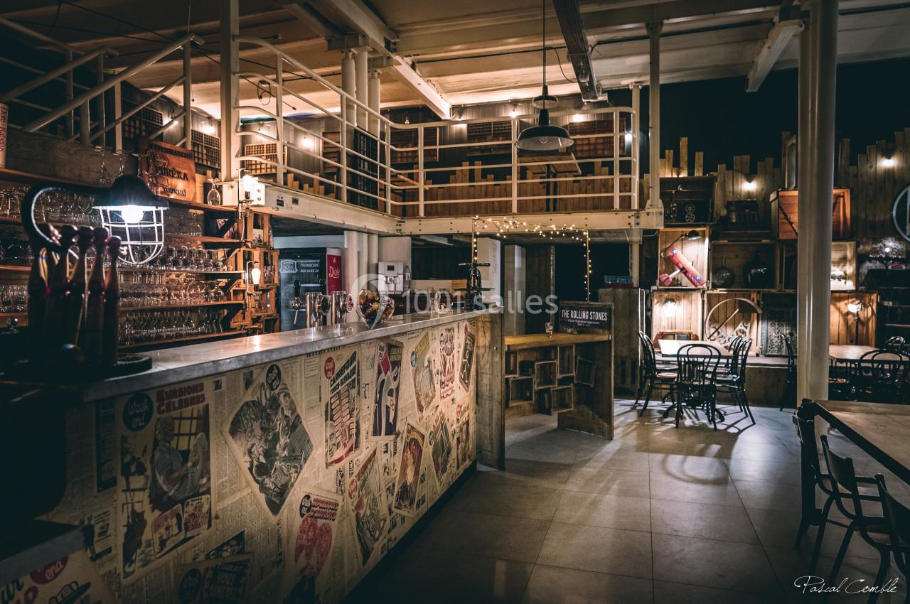 Intérieur d'un bar au style industriel avec comptoir décoré de journaux, tables et éclairage tamisé.