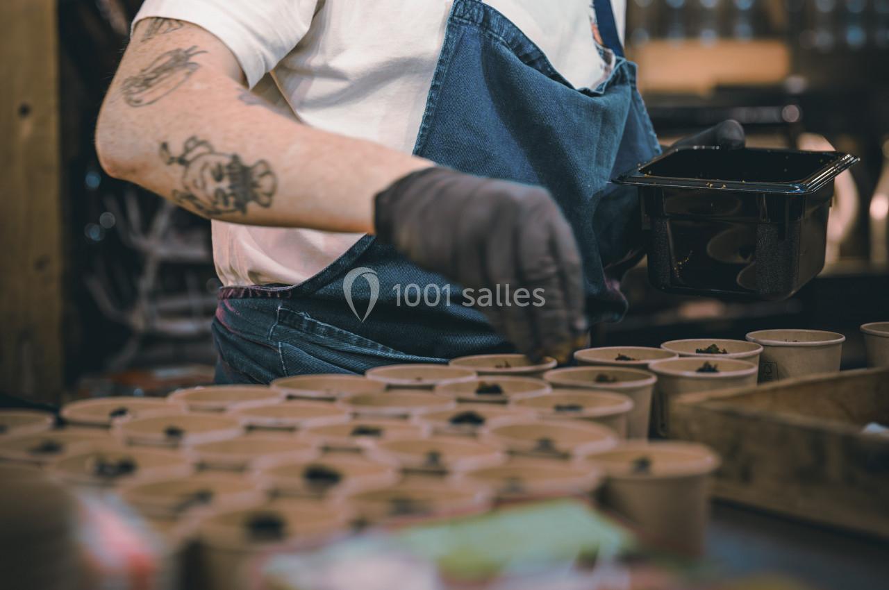 Une personne en tablier bleu remplit des pots en carton avec de la terre dans un environnement de travail.