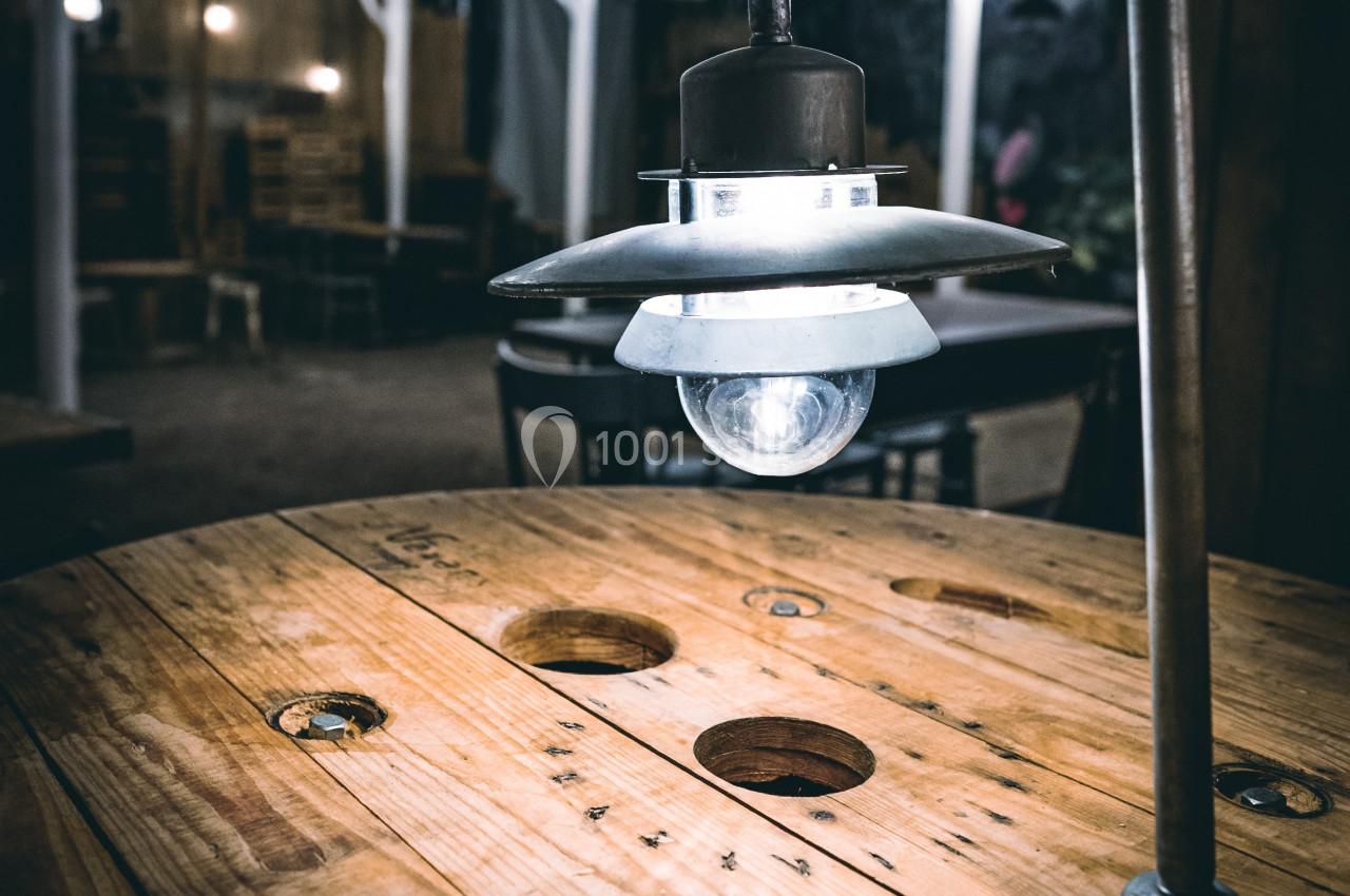 Suspension lumineuse en métal éclairant une table ronde en bois avec des trous, dans un espace intérieur sombre.