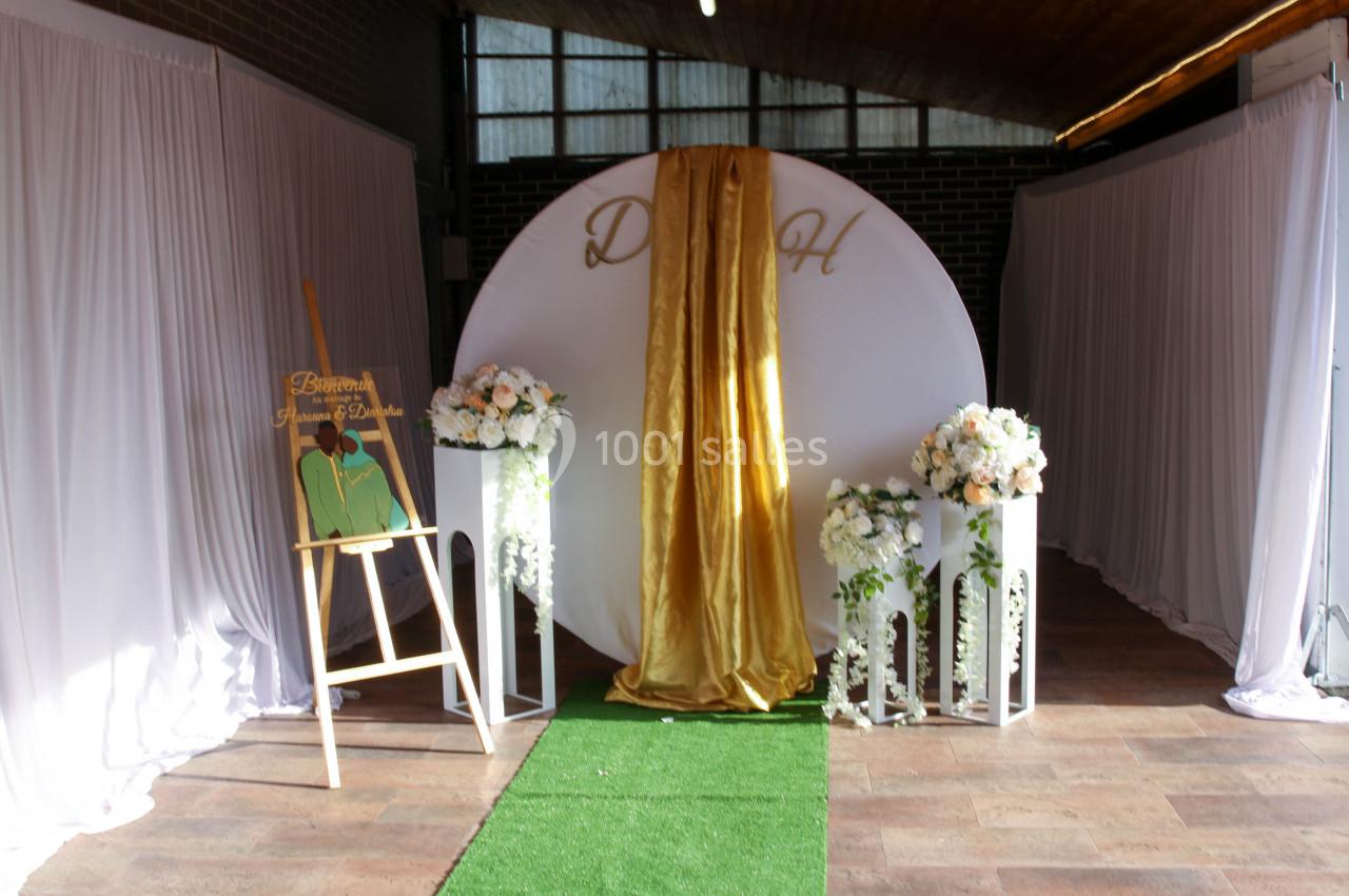 Décor de cérémonie avec un fond circulaire blanc, drapé doré, fleurs blanches et un chevalet de bienvenue.