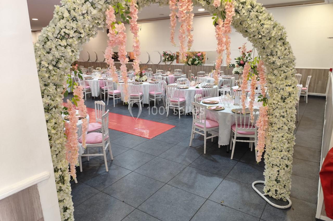 Arche florale blanche et rose menant à une salle de réception décorée avec des tables dressées et des chaises blanches.