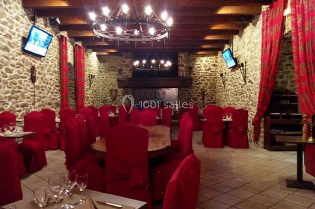 Salle de restaurant avec murs en pierre, chaises rouges, tables dressées et lustres suspendus au plafond en bois.