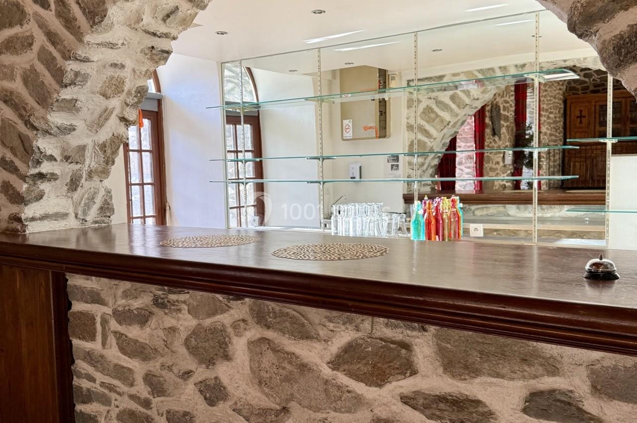 Comptoir en bois avec murs en pierre, étagères en verre, bouteilles colorées et lumière naturelle entrant par une porte…