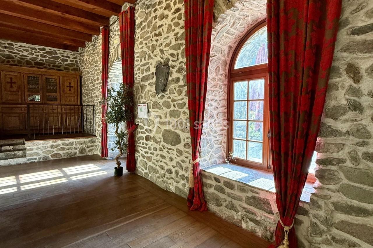 Mur en pierre avec une grande fenêtre cintrée, encadrée de rideaux rouges, éclairant une pièce au sol en bois.