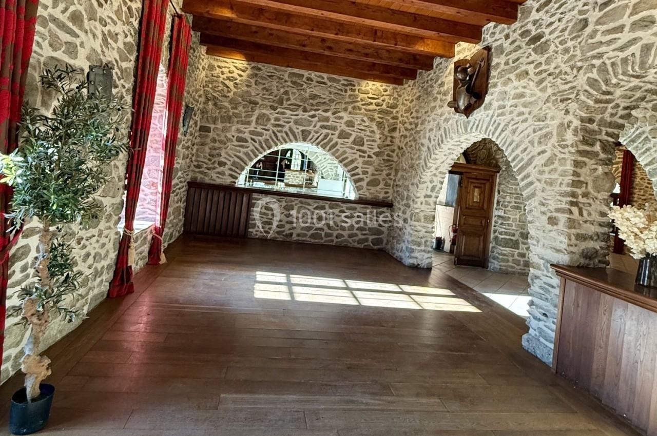 Salle en pierre avec poutres apparentes, grandes fenêtres voûtées, parquet en bois et décoration rustique.