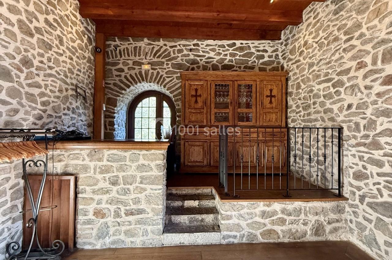 Mur en pierre apparente, escalier en pierre, meuble en bois sculpté et porte voûtée avec lumière naturelle.