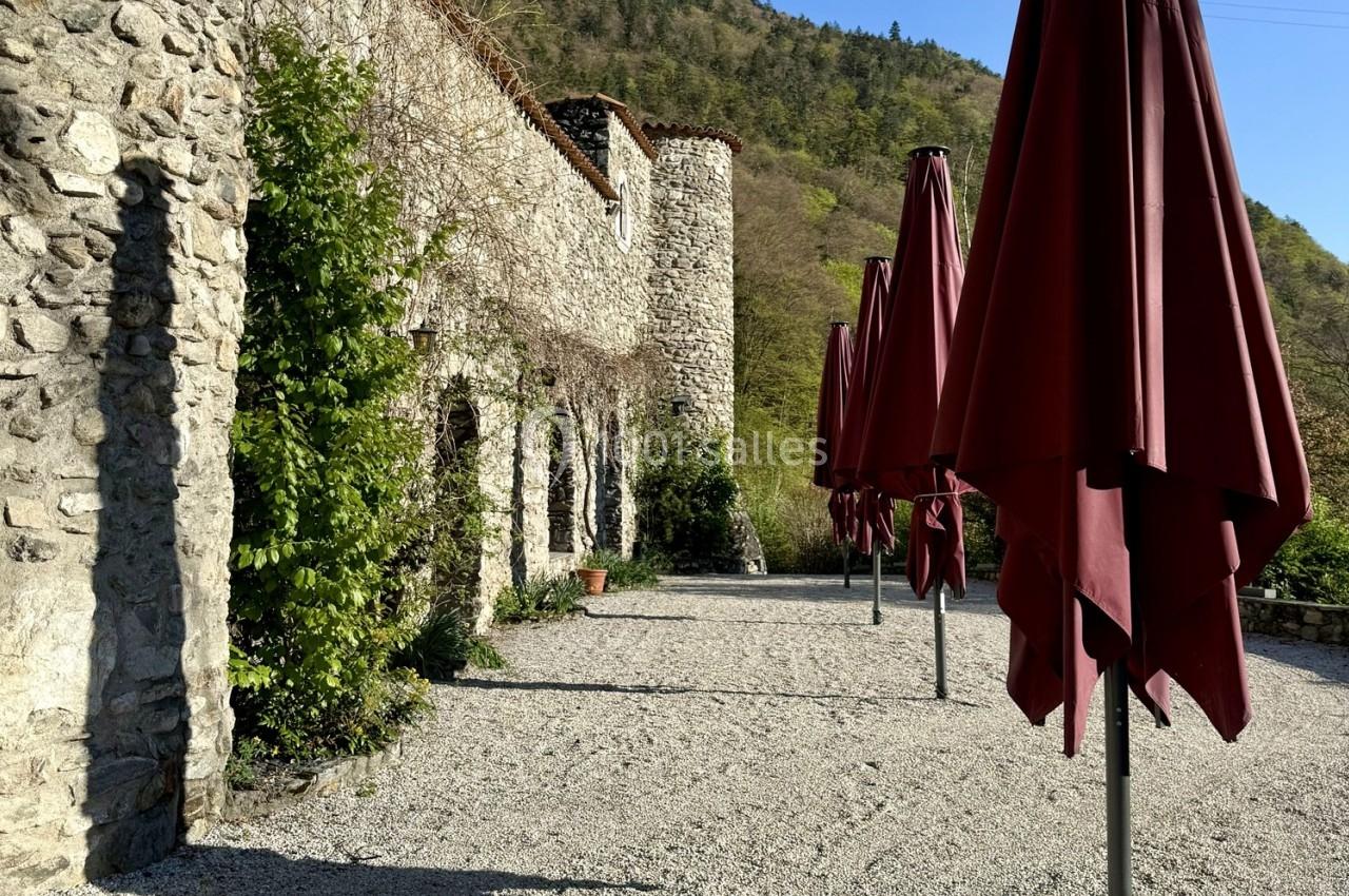 Cour extérieure avec murs en pierre, parasols rouges fermés et végétation en arrière-plan.