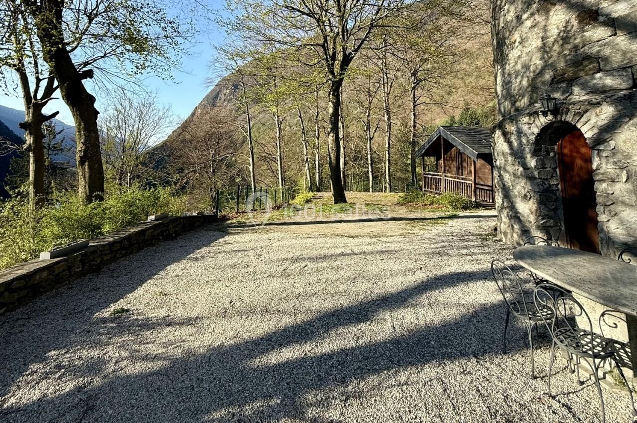 Cour en gravier bordée d'arbres, avec une table en fer forgé, une maison en pierre et un chalet en bois en arrière-plan.