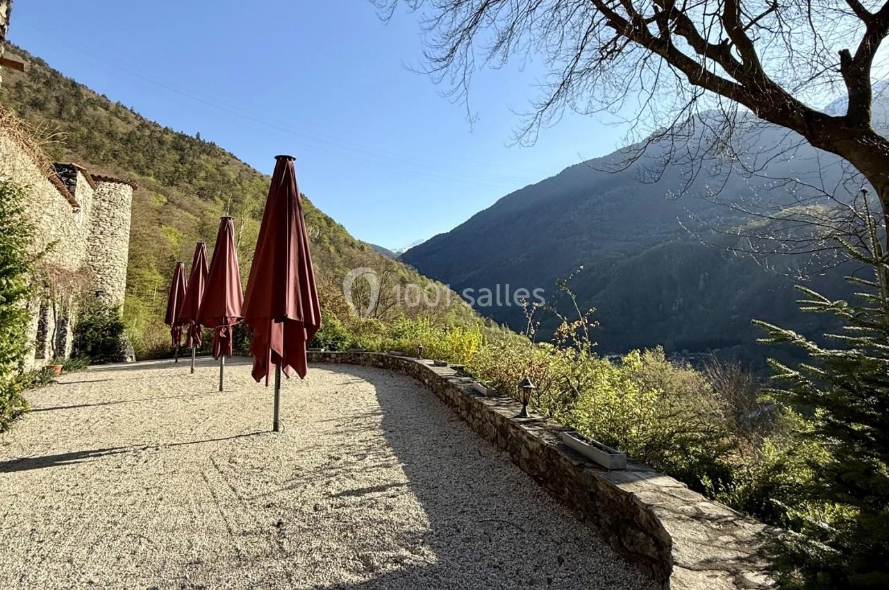 Terrasse en gravier avec parasols rouges fermés, bordée de végétation, offrant une vue sur des montagnes ensoleillées.