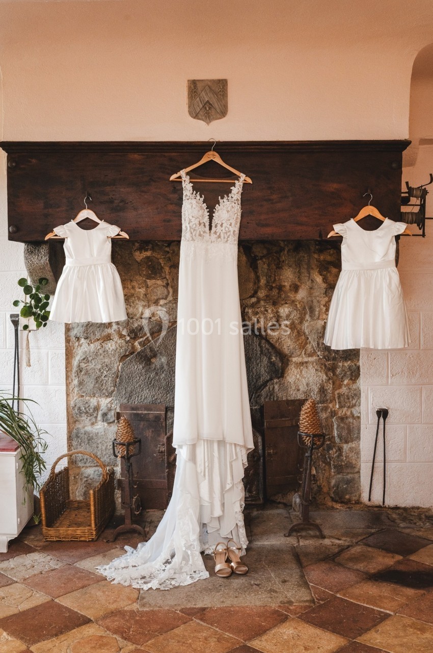 Robe de mariée blanche et deux robes d'enfant suspendues devant une cheminée en pierre dans une pièce rustique.