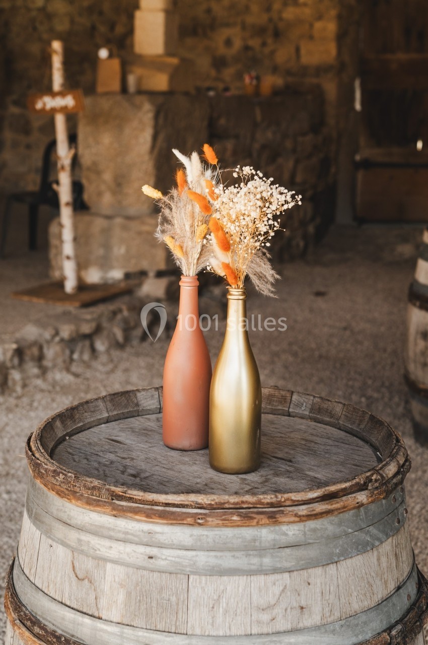 Deux bouteilles décoratives avec des fleurs séchées posées sur un tonneau en bois dans un espace rustique.