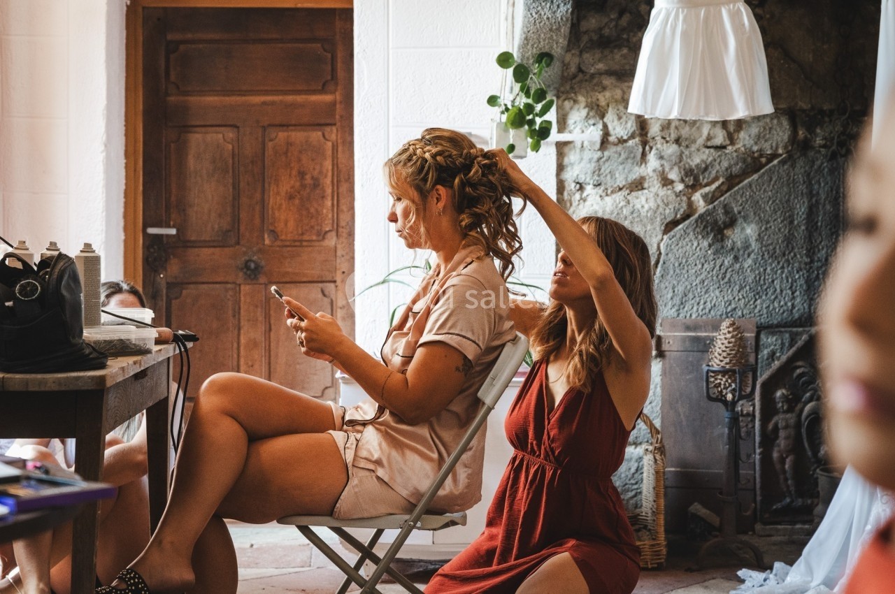 Une femme assise consulte son téléphone pendant qu'une autre lui coiffe les cheveux dans une pièce rustique.
